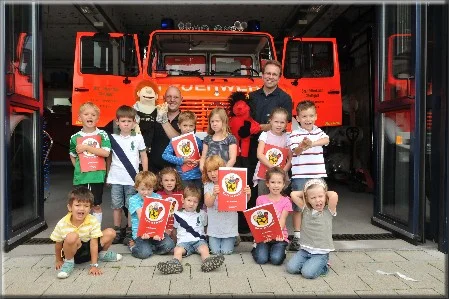 Kindergruppe posiert vor dem geöffneten Tor eines Feuerwehrhauses mit einem Feuerwehrfahrzeug im Hintergrund